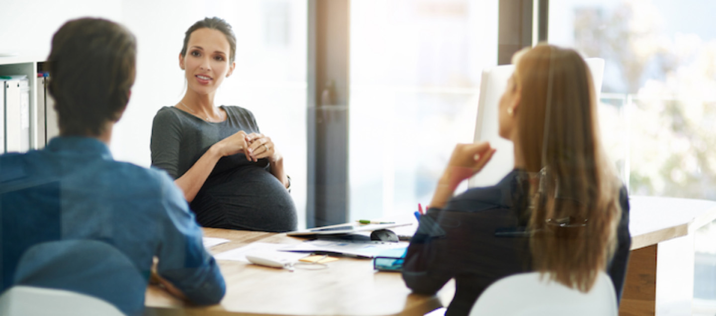 A pregnant woman in a grey dress is seated at a meeting table, engaged in conversation with two people. The setting is a modern, bright office with large windows, and various documents and a smartphone are visible on the table.