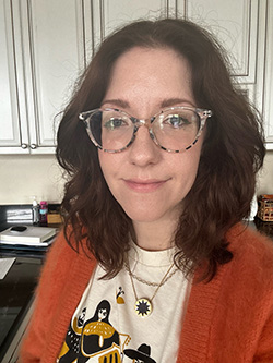 Woman with shoulder-length brown hair and clear-framed glasses is smiling while wearing a graphic tee with a design and an orange cardigan.