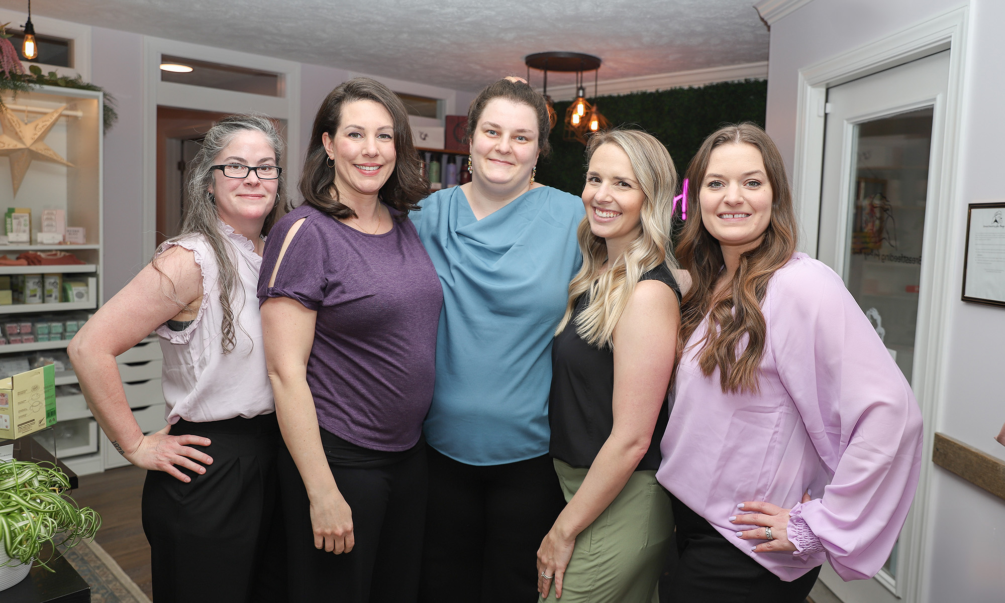 Five smiling women standing indoors, from left to right: a woman with glasses in a pink blouse, a woman in a purple blouse, a woman with tied-back hair in a blue top, a woman in a black top, and a woman in a purple blouse.