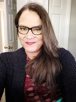 Woman with long brown hair and glasses is smiling while wearing a patterned red top and a dark cardigan.