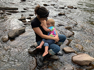 A woman in a black shirt and jeans is sitting on rocks in a shallow stream, breastfeeding a child in a colorful tie-dye shirt. The stream is surrounded by smooth rocks and flowing water.