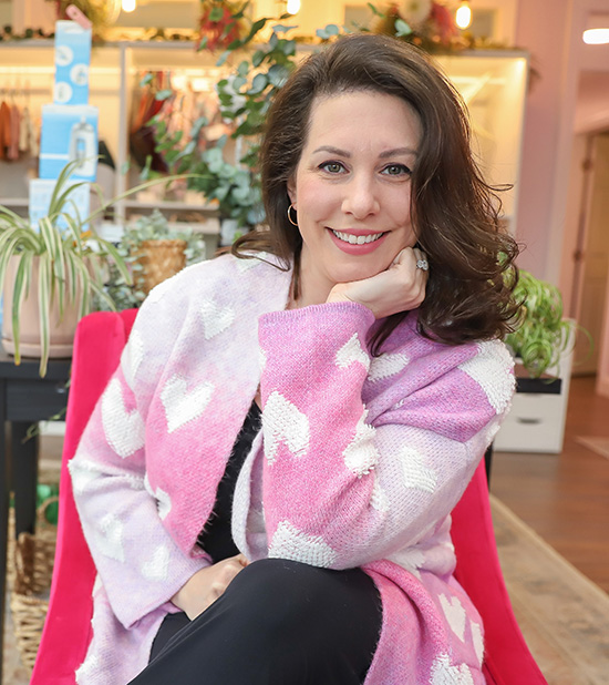 Woman seated comfortably and resting her chin in the palm of her hand. She is wearing a pink sweater with white heart shapes on it.