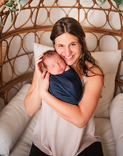 A smiling woman is seated and holding a sleeping baby swaddled in a dark blue blanket.