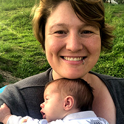 Woman with short brown hair is smiling while holding a baby against their chest, posing outdoors with green grass in the background.