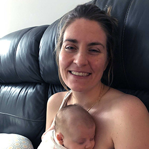 A smiling woman is sitting on a black leather couch, holding a sleeping baby close to her chest.